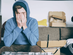 A person with their hoodie up, seated on a couch and surrounded by cardboard boxes. They look distressed.